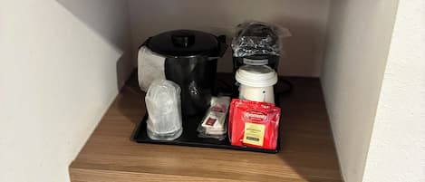 Un área de cocina con un microondas en la repisa superior, una cafetera, un hervidor y un dispensador de cápsulas de café en la repisa central, y un electrodoméstico negro en la repisa inferior.