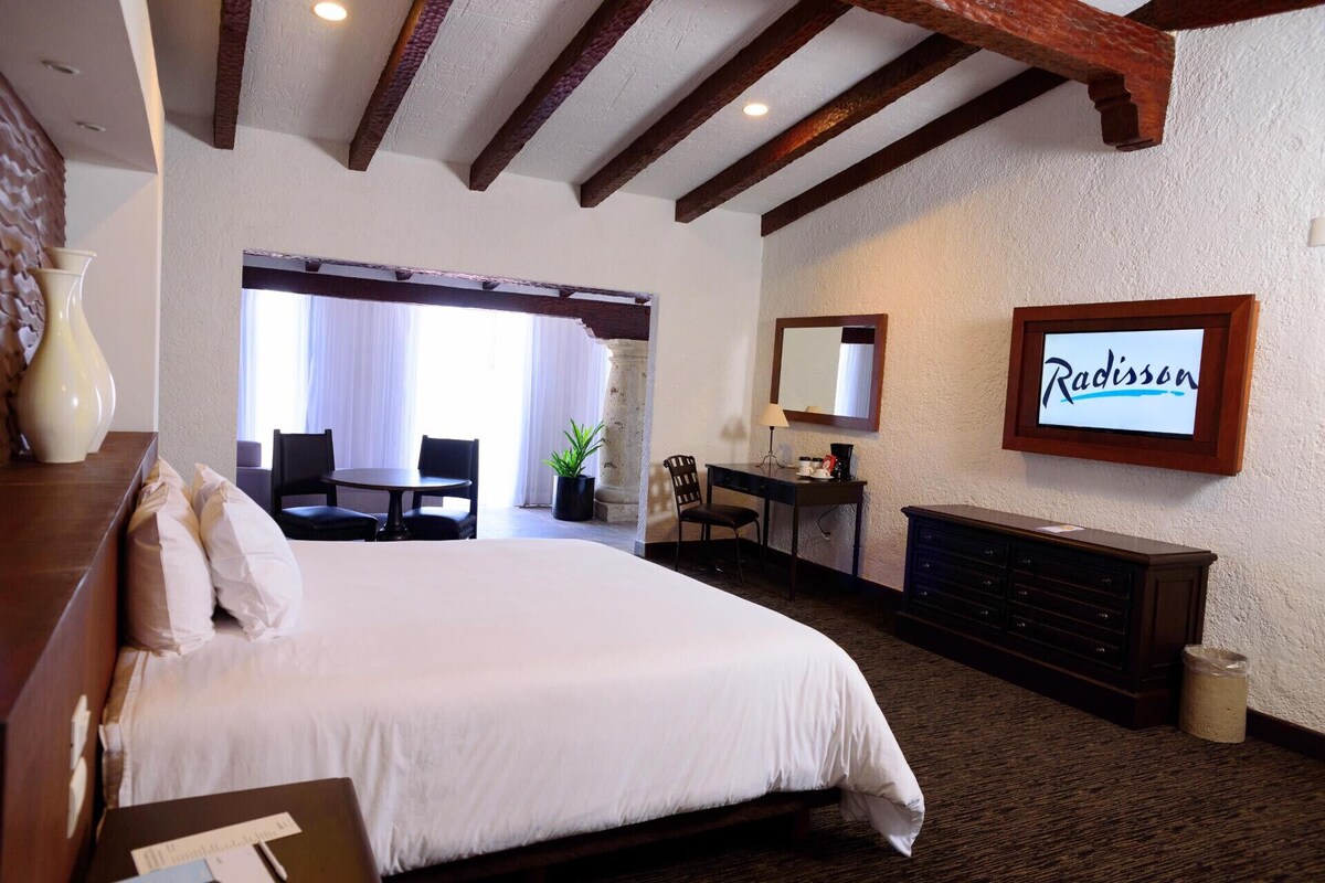 A hotel room with a large bed, a desk, a chair, a dresser, a mirror, and a sign that reads 'Radisson.'