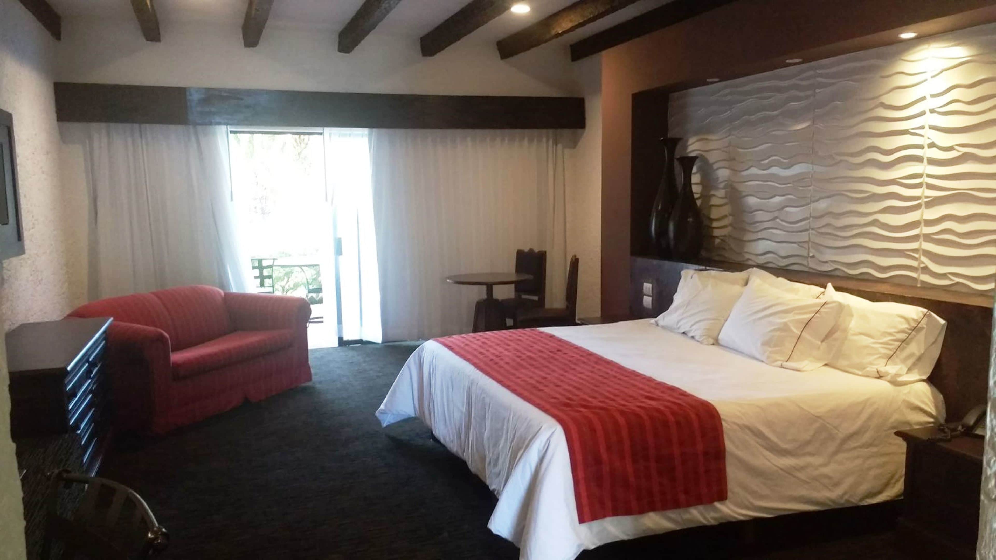 A hotel room with a bed, a red armchair, a small table, and a desk.