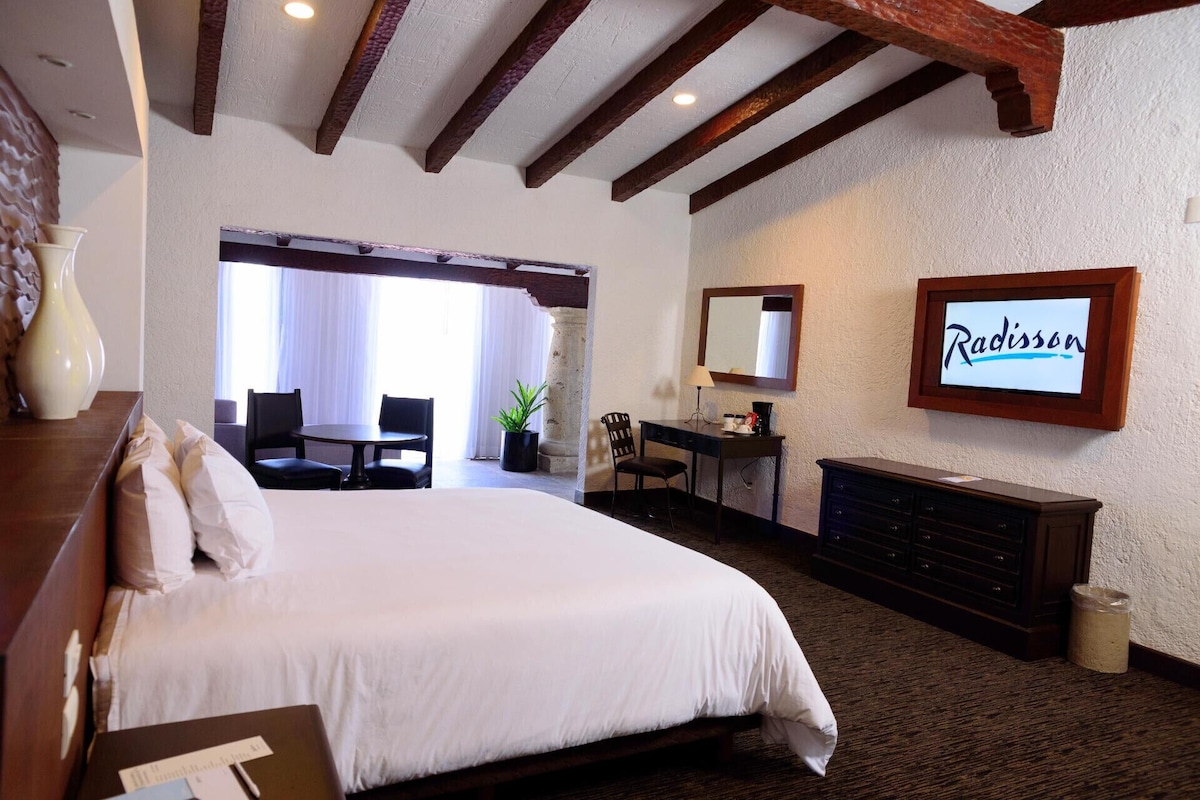 A hotel room with a large bed, a desk, a chair, a dresser, a mirror, and a sign that reads 'Radisson.'