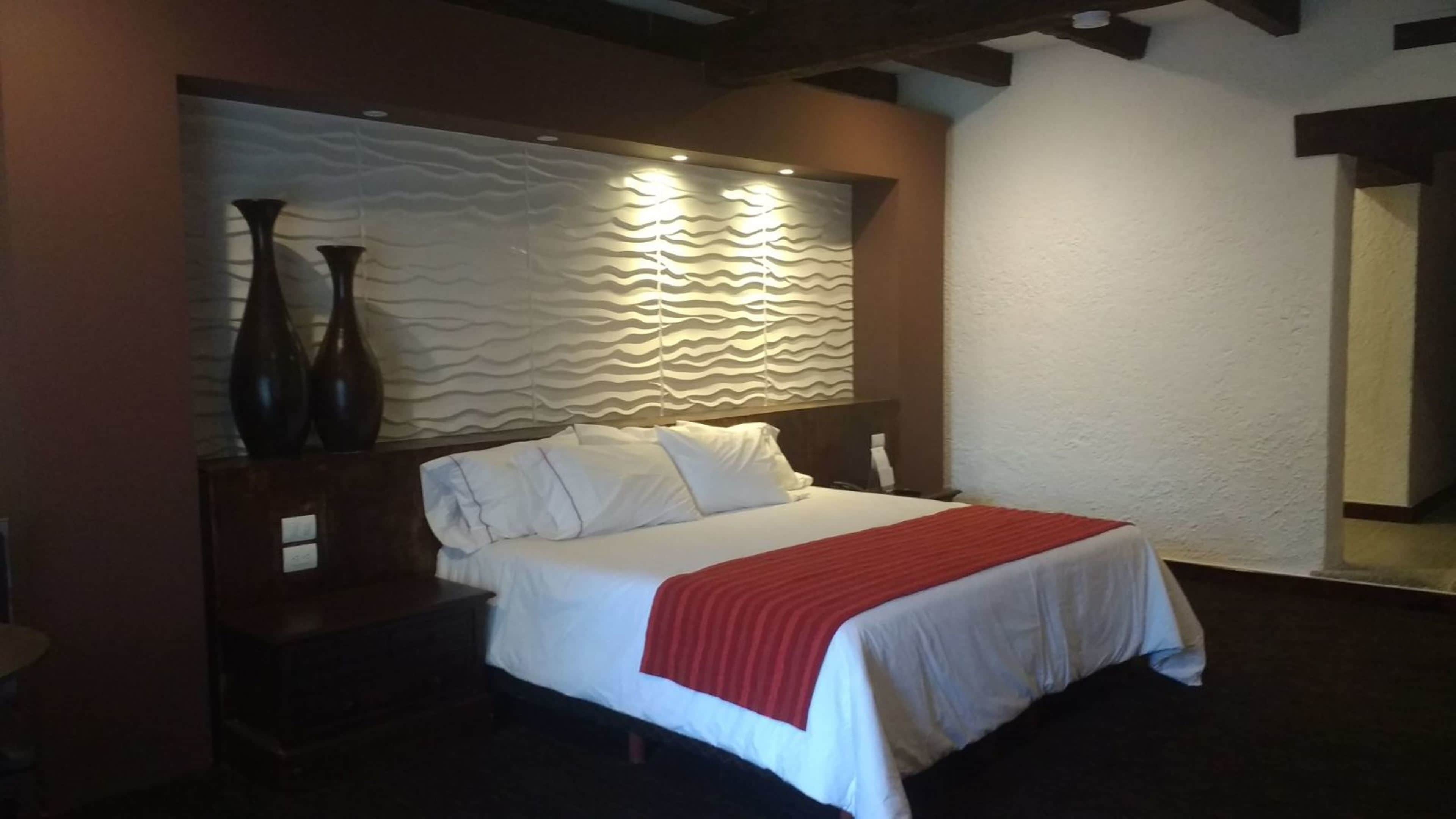 Una cama bien hecha con sábanas blancas y una manta roja, una mesita de noche de madera con un jarrón, y una pared con textura detrás de la cama.