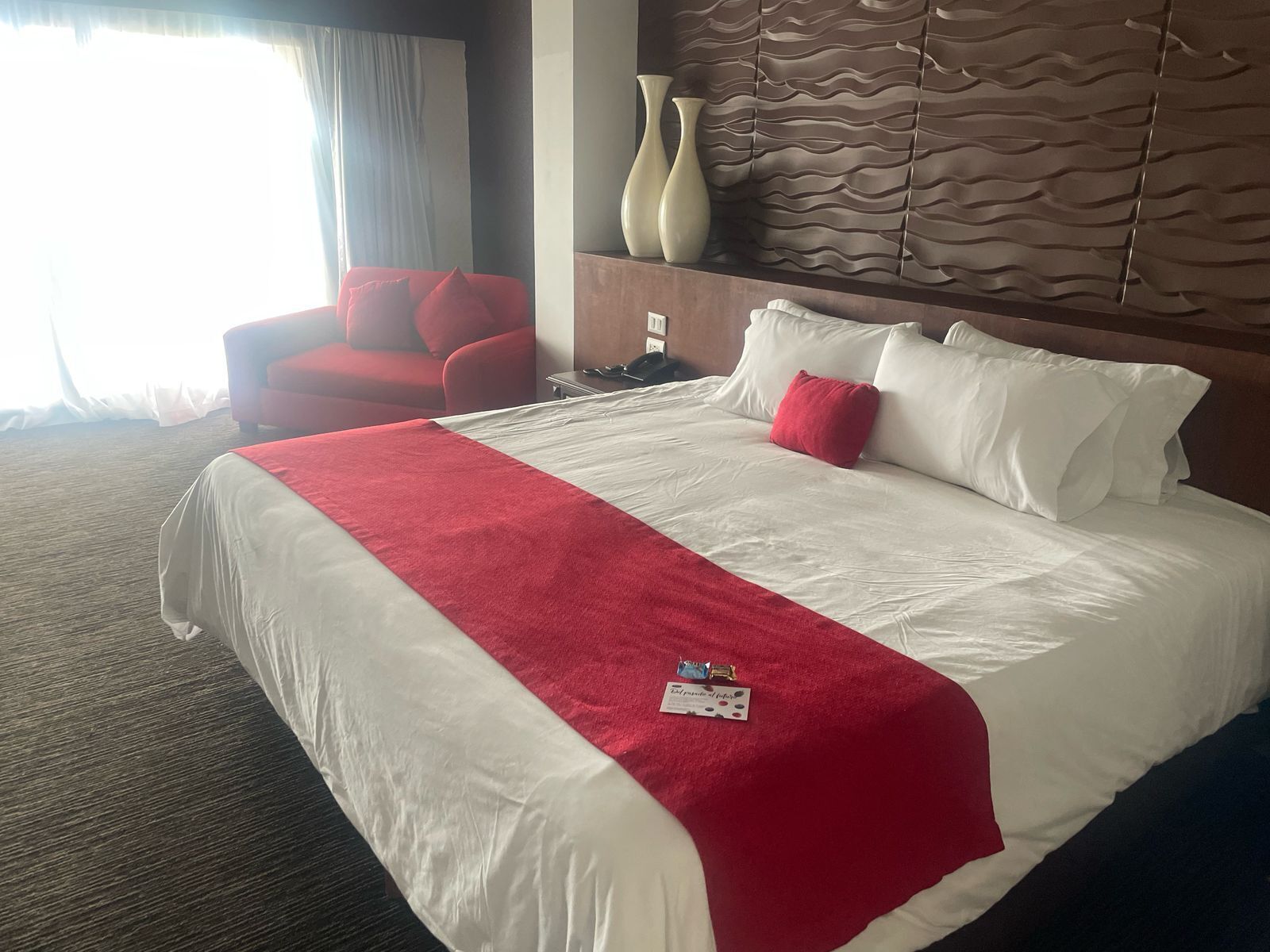 A hotel room with a large bed, red and white bedding, a red armchair, and a decorative wall panel.