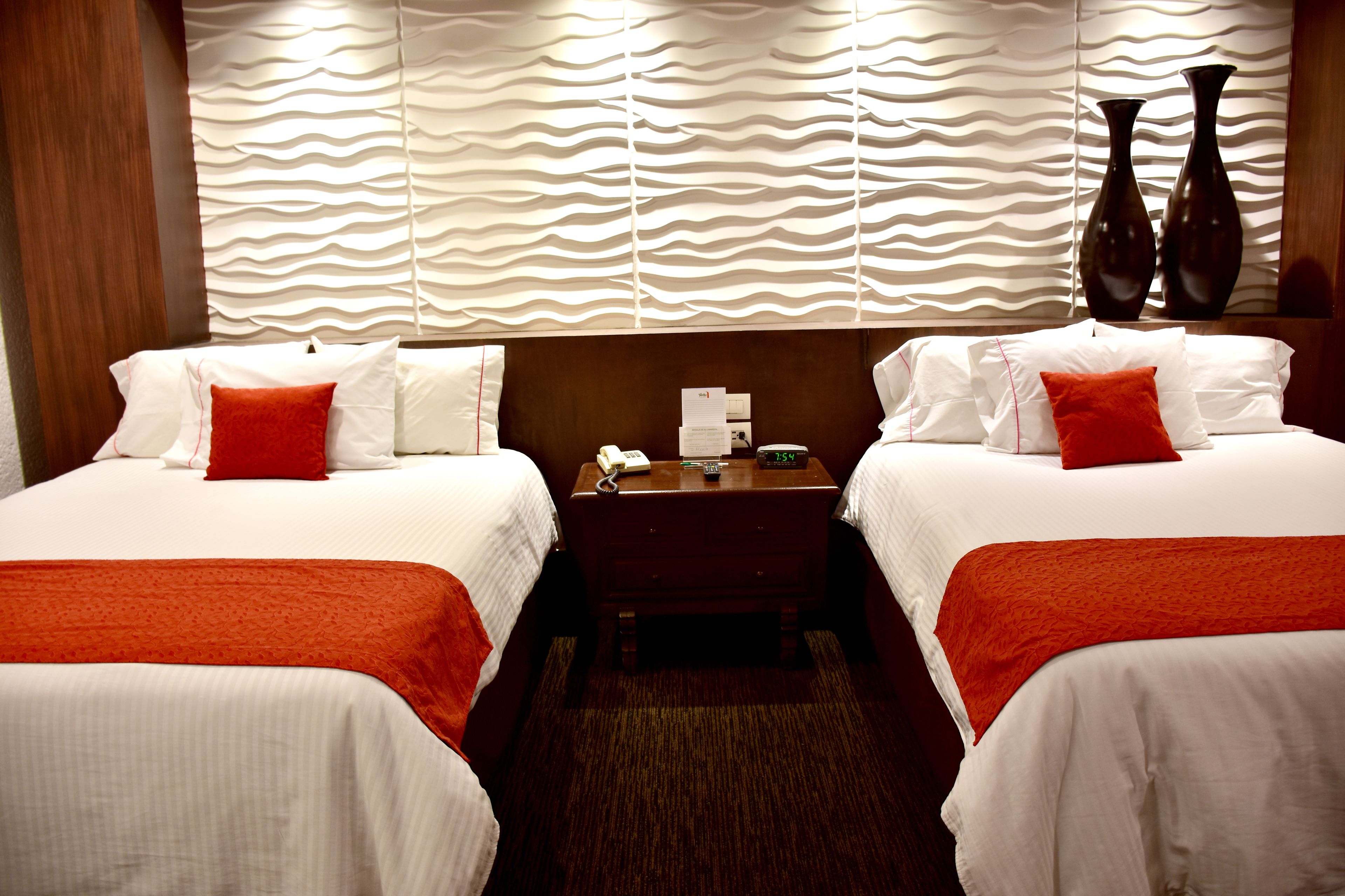 A hotel room with two beds, each with white linens and red accents, a wooden headboard, a nightstand with a phone and clock, and a textured wall behind the beds.