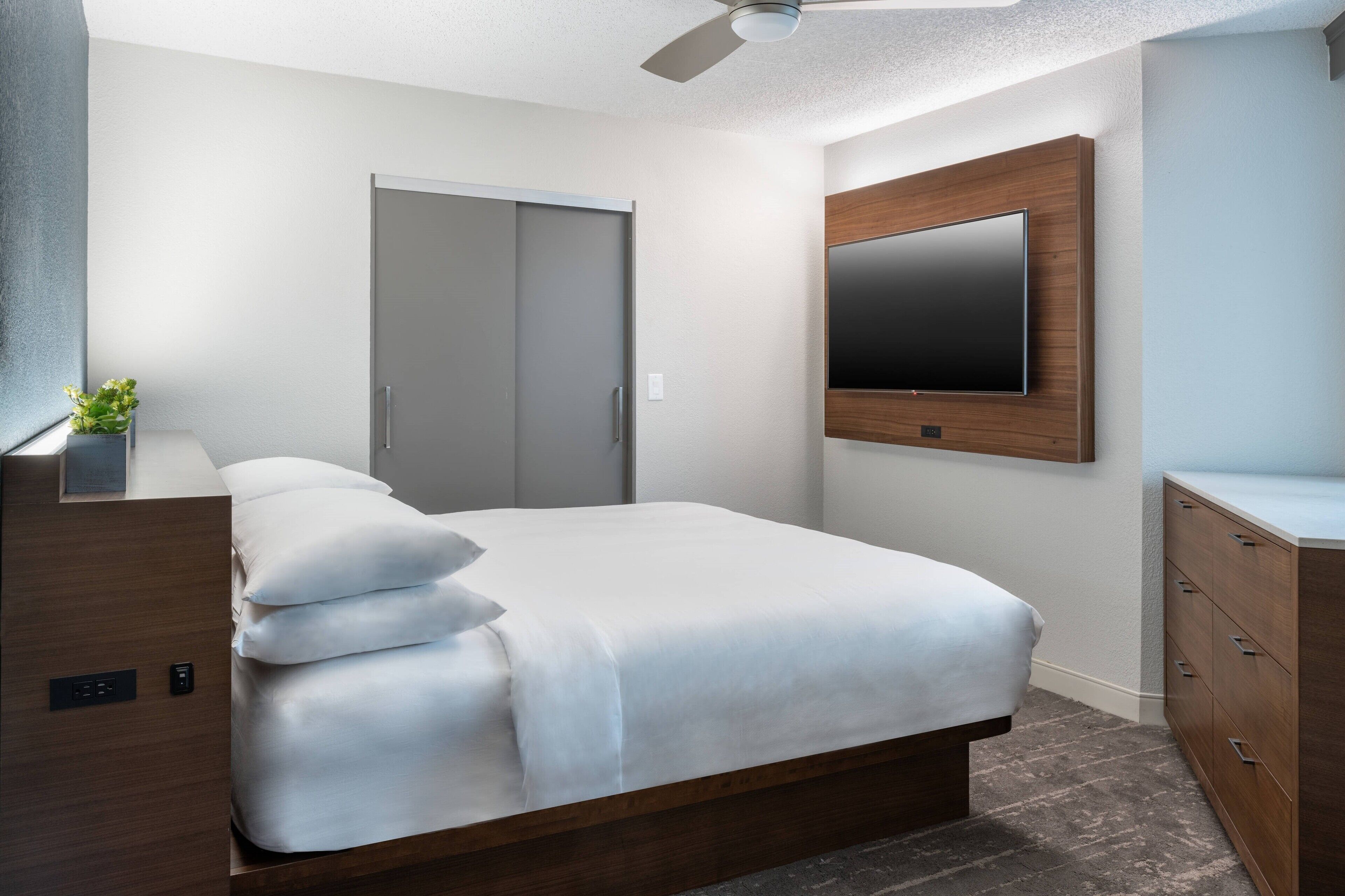 Une chambre moderne avec un lit, un téléviseur fixé au mur, un ventilateur de plafond et une armoire.