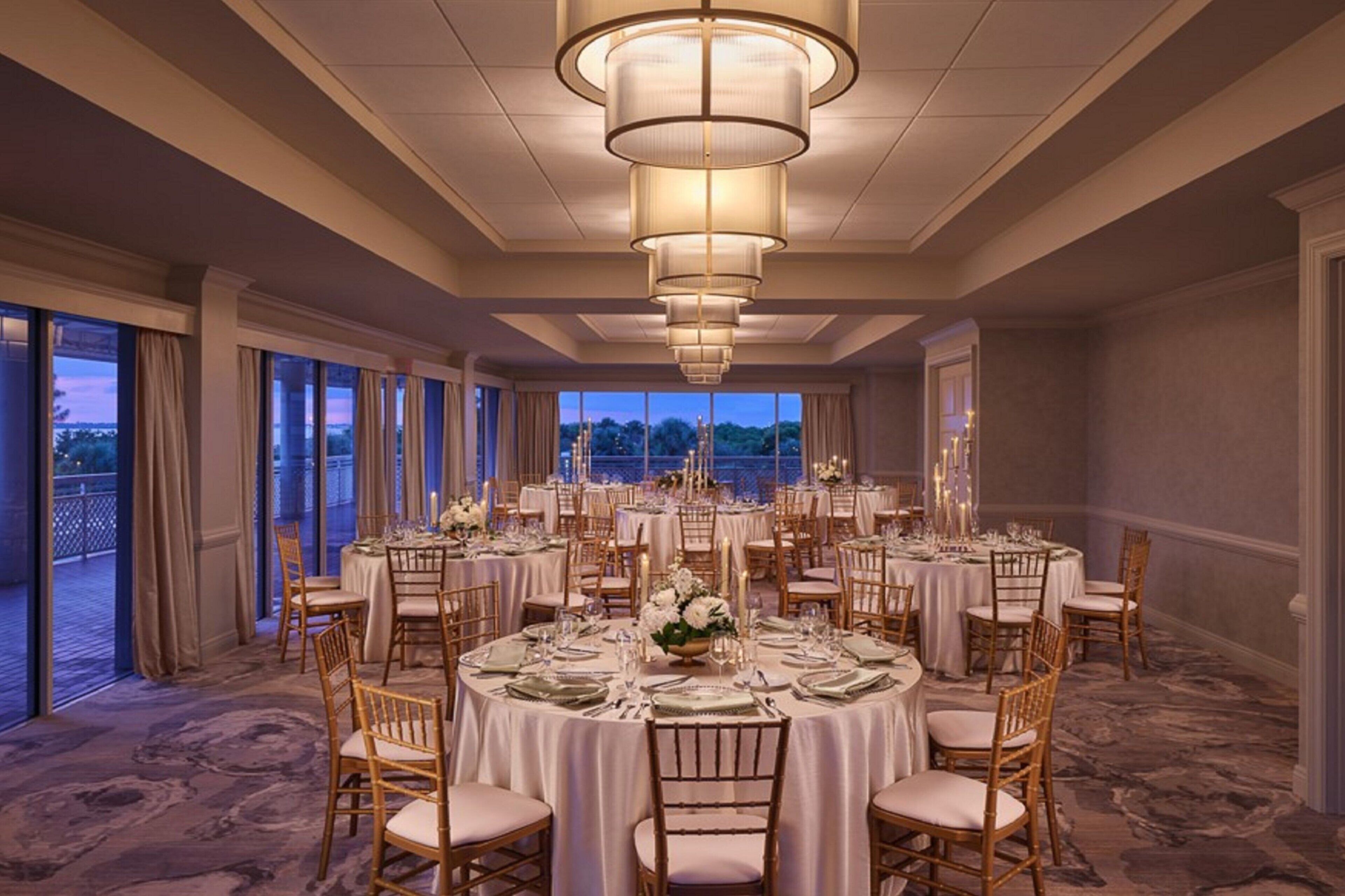 A banquet hall with round tables set for dining, large chandeliers, and a view of the outdoors.