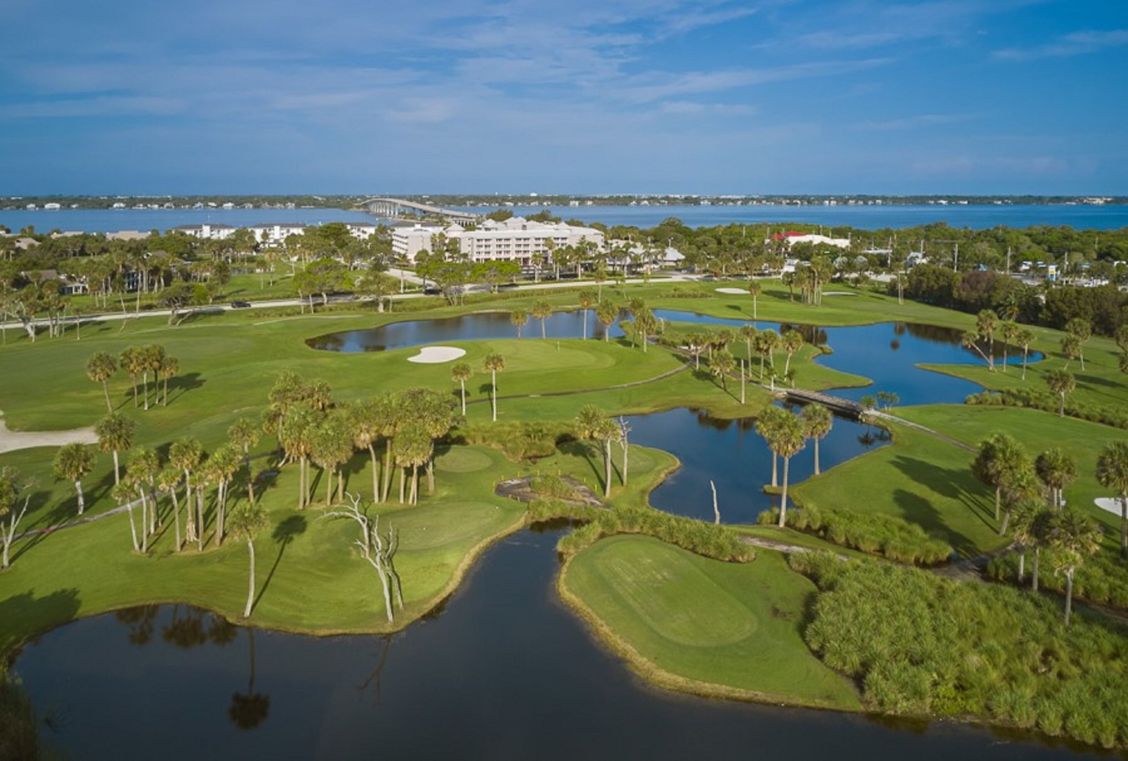 A golf course with water hazards, palm trees, and a large building in the background.