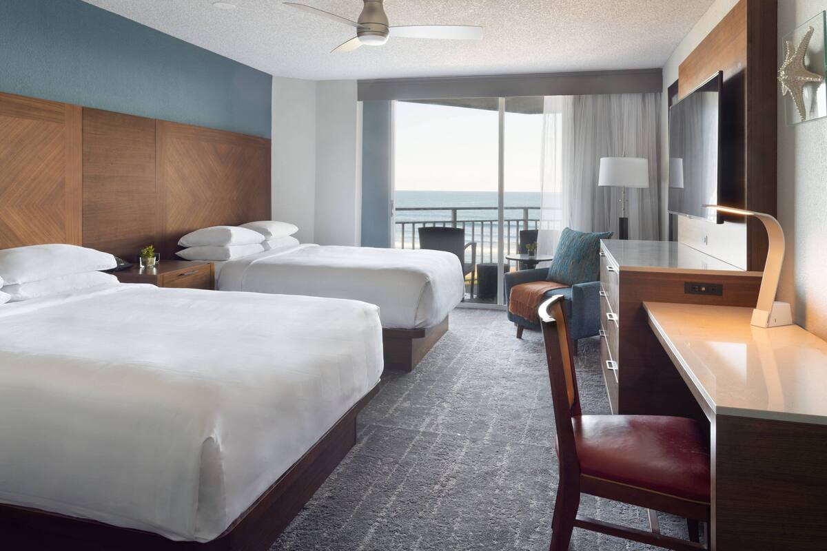 Habitación de hotel con dos camas, un escritorio, una silla, un televisor y un balcón con vista al mar.
