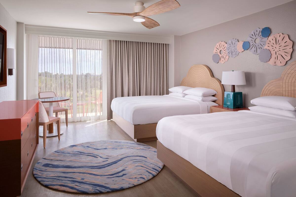 Habitación de hotel con dos camas, un escritorio, una silla, un ventilador de techo y un ventanal grande con cortinas.