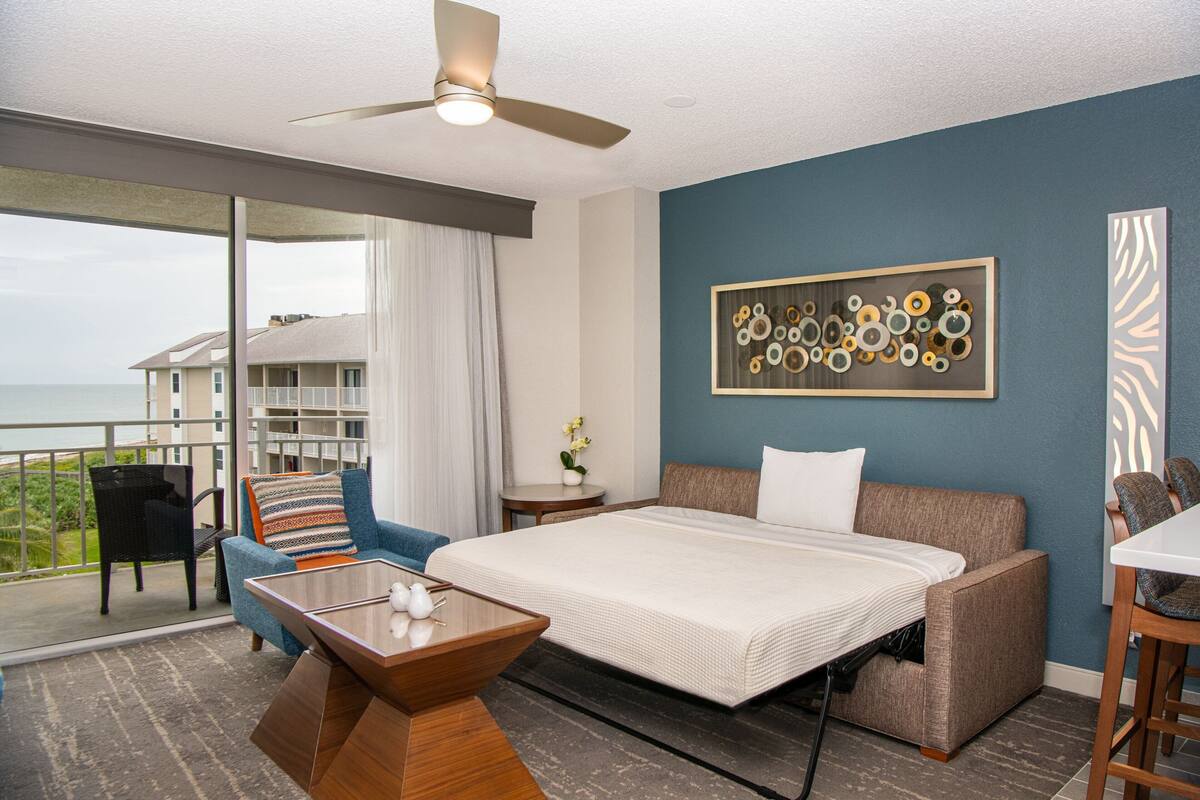 Habitación de hotel con cama, sofá, mesa de comedor y balcón con vista al mar.