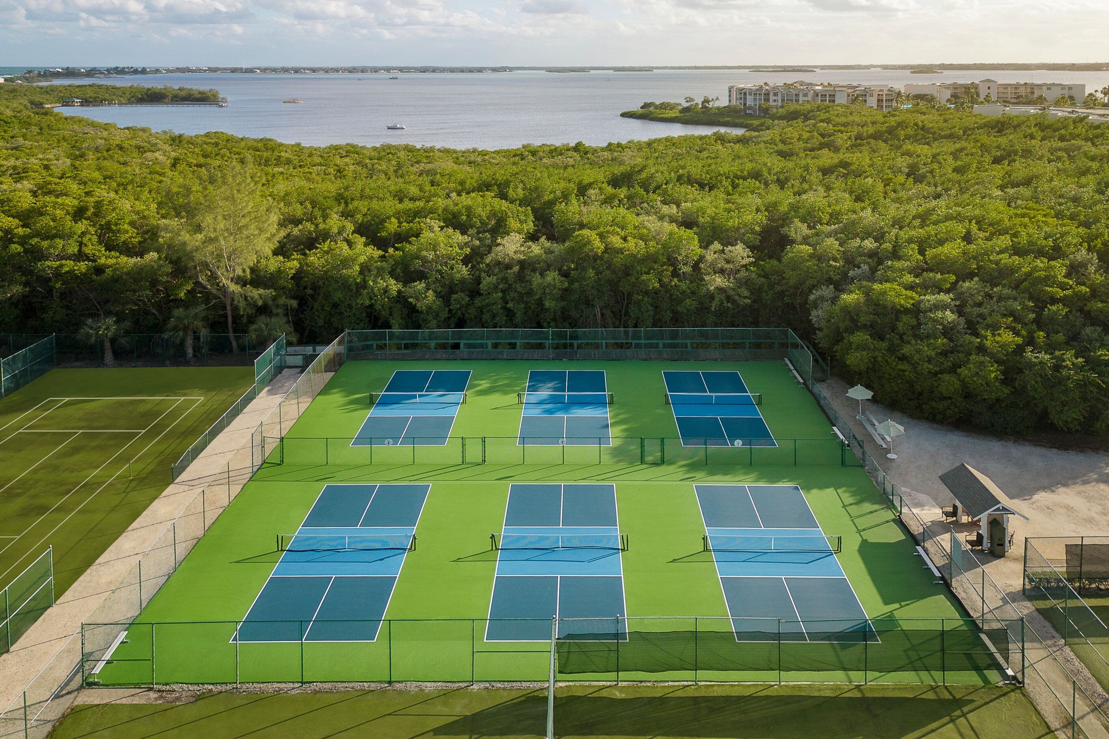 Un complexe de courts de tennis comprenant plusieurs terrains, entouré d’une végétation luxuriante et avec un plan d’eau en arrière-plan.