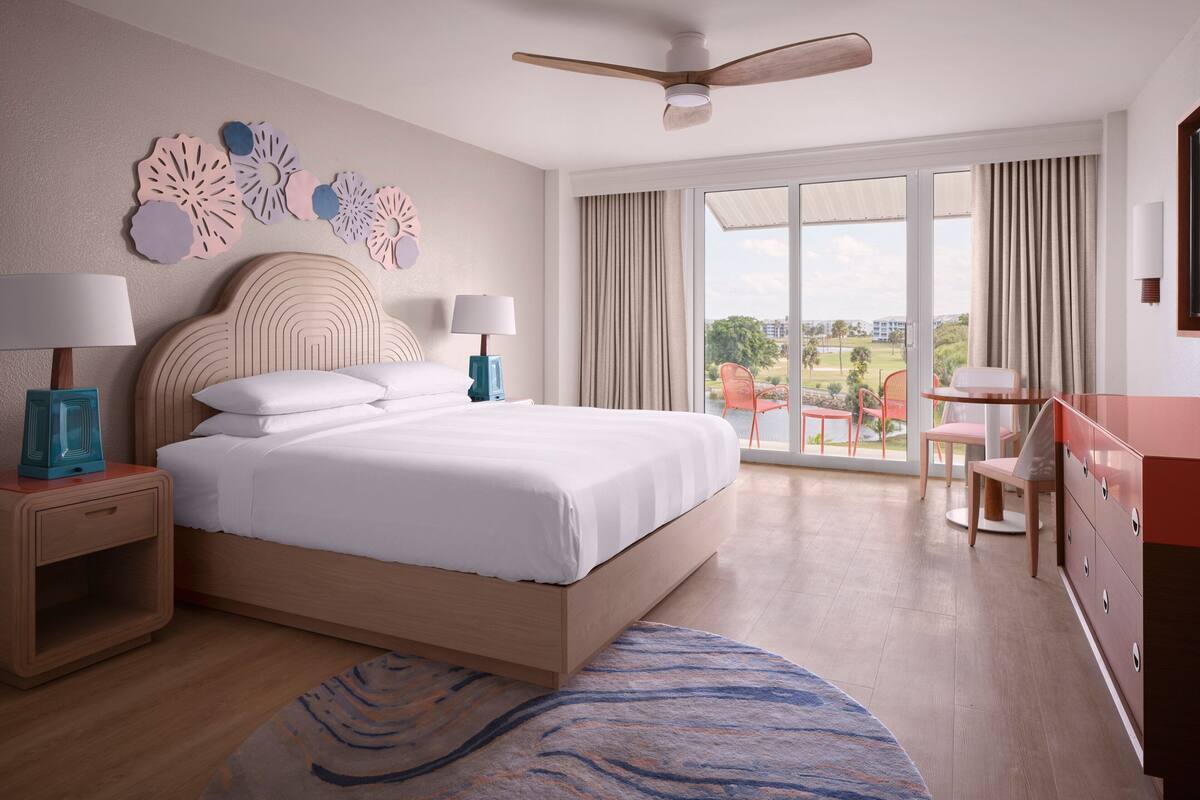 Habitación de hotel con una cama grande, dos mesitas de noche con lámparas, un ventilador de techo, vistas al exterior a través de un ventanal corredero y una pequeña mesa con sillas.