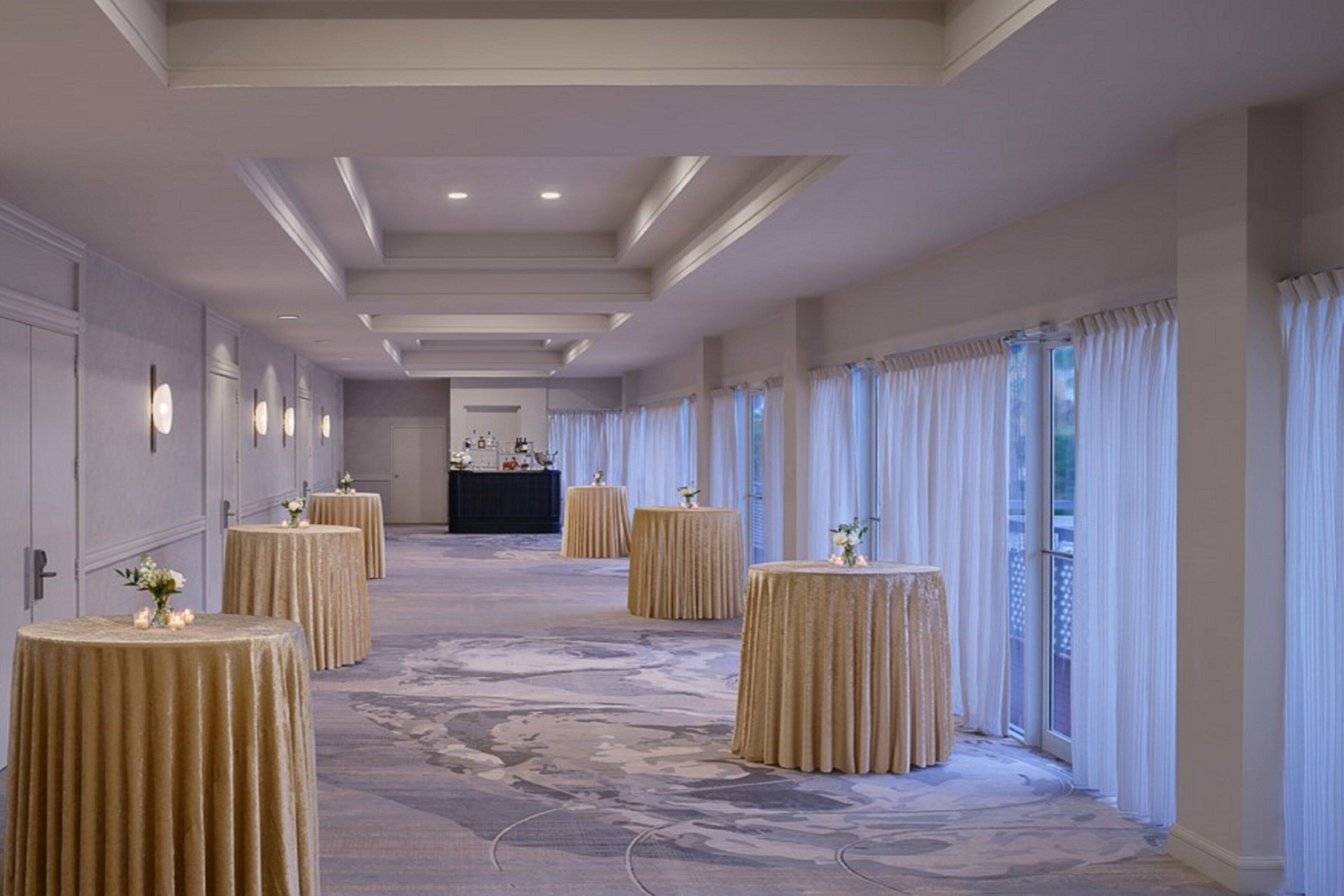 A hotel corridor with round tables, a reception desk, and large windows with curtains.