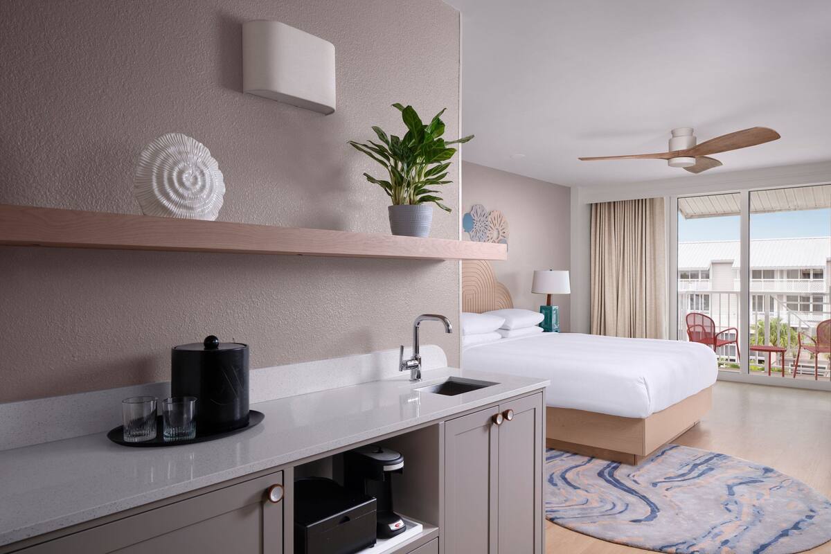 Una habitación de hotel moderna con una cama grande, un escritorio integrado con cafetera, un lavamanos pequeño, ventilador de techo y balcón con mobiliario exterior.