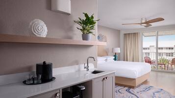 Una habitación de hotel moderna con una cama grande, un escritorio integrado con cafetera, un lavamanos pequeño, ventilador de techo y balcón con mobiliario exterior.
