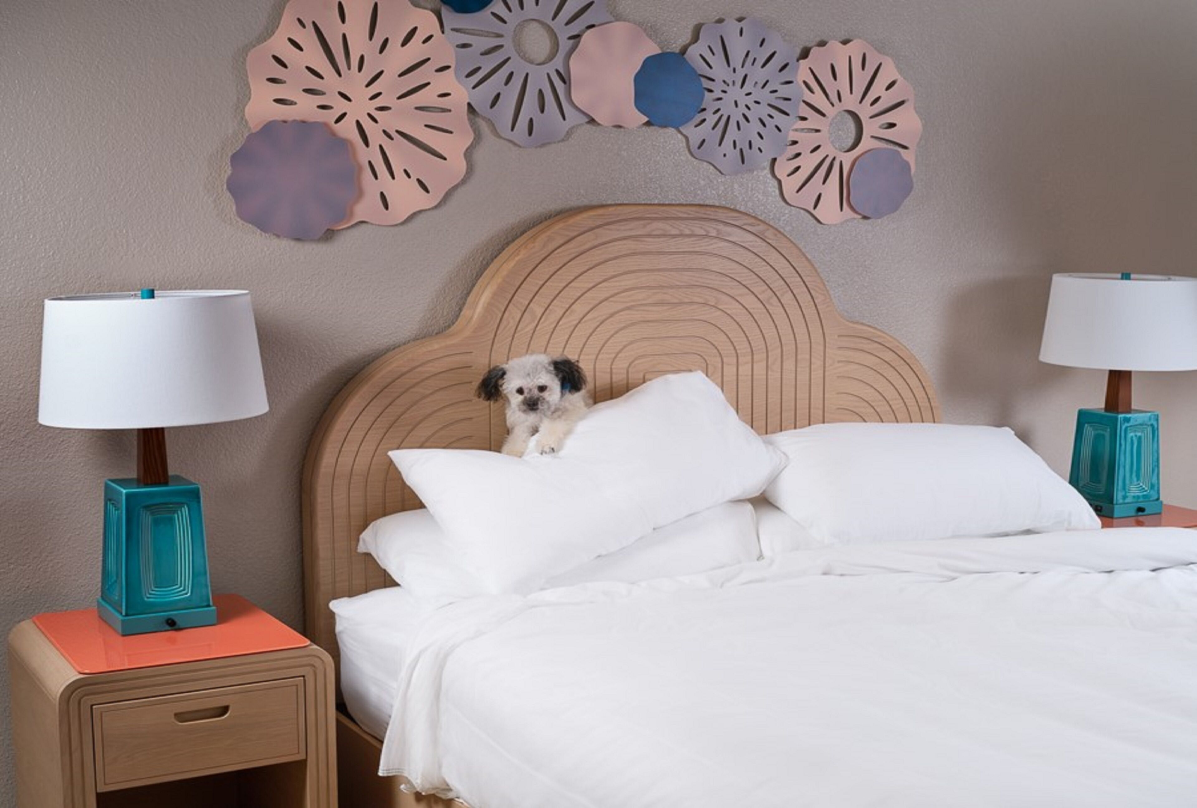 A hotel room with a bed, two bedside tables with lamps, a decorative wall piece, and a small dog on the bed.