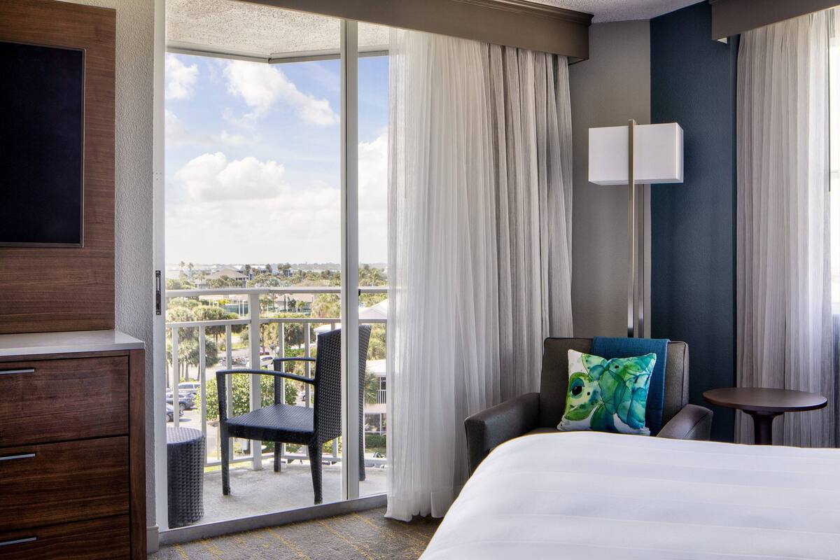 Habitación de hotel con una cama grande, un escritorio, una silla, una mesita y vistas a la ciudad a través de amplias ventanas.