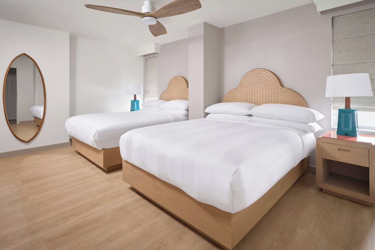 Habitación de hotel con dos camas, ventilador de techo, espejo y mesita de noche con lámpara.