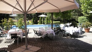 An outdoor patio with tables and chairs, a pool in the background, and a large umbrella providing shade.