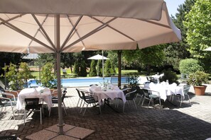 An outdoor patio with tables and chairs, a pool in the background, and a large umbrella providing shade.