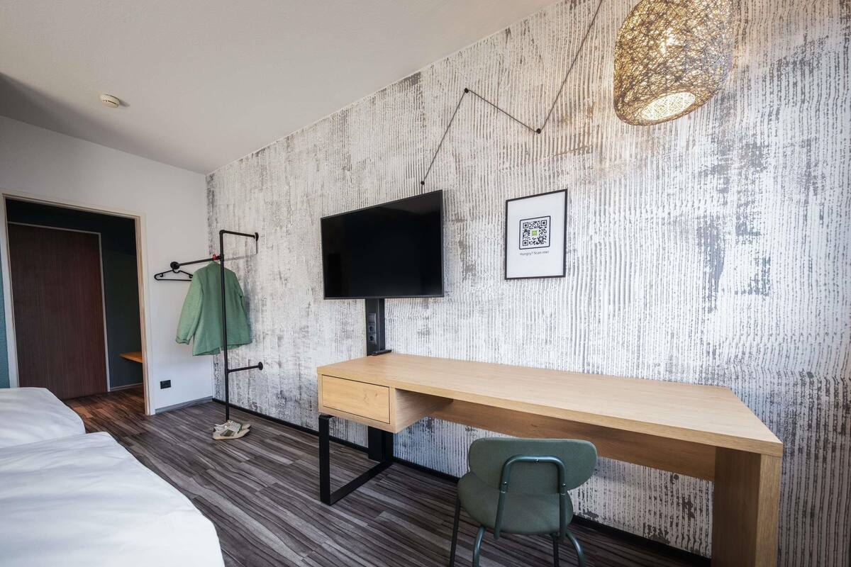 A modern hotel room with a wooden desk, a green chair, a flat-screen TV mounted on the wall, and a QR code for digital services.