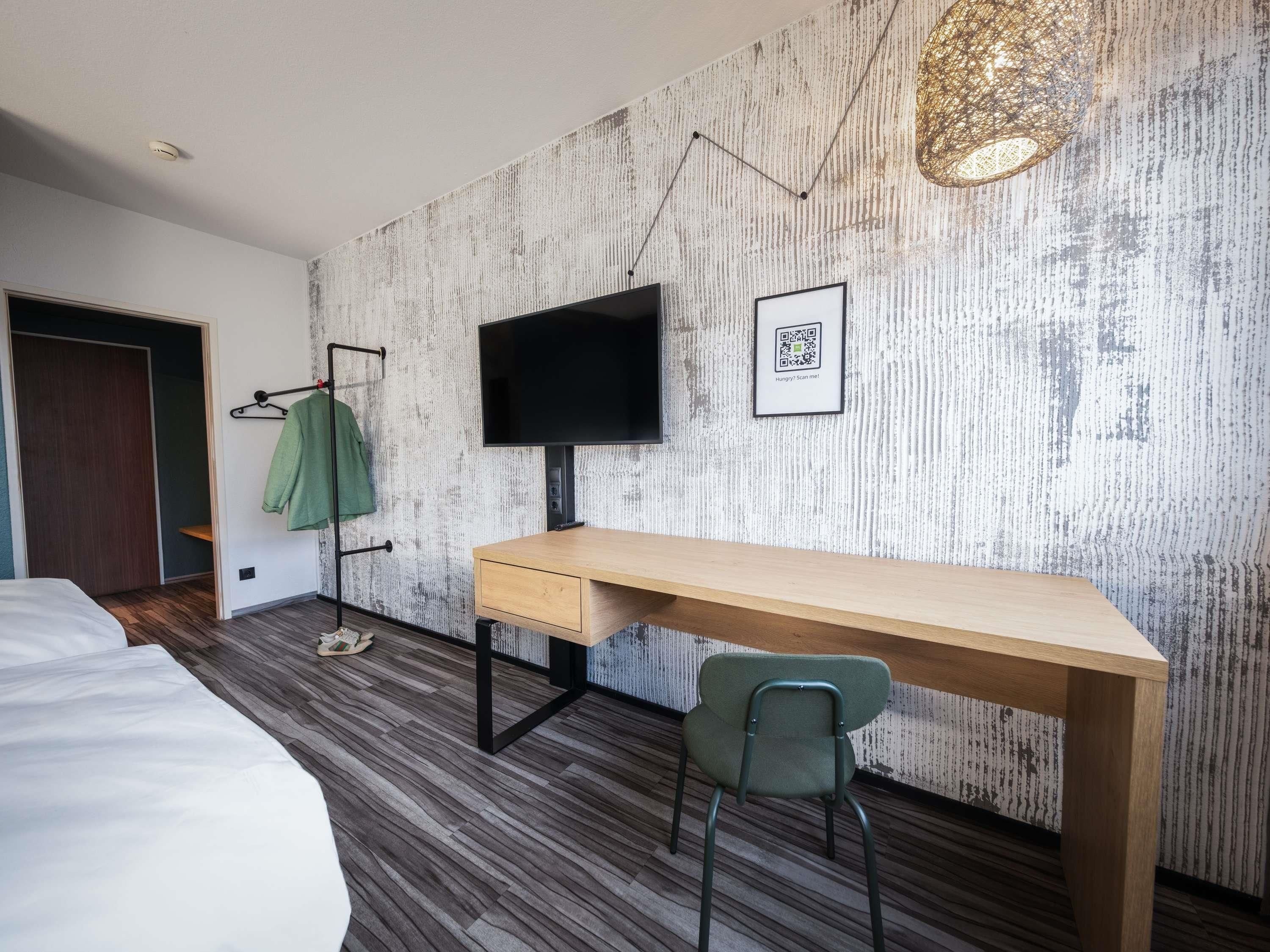 Ein modernes Hotelzimmer mit Schreibtisch aus Holz, einem grünen Sessel, einem flachbildschirmfernseher an der Wand und einem QR-Code für digitale Dienstleistungen.