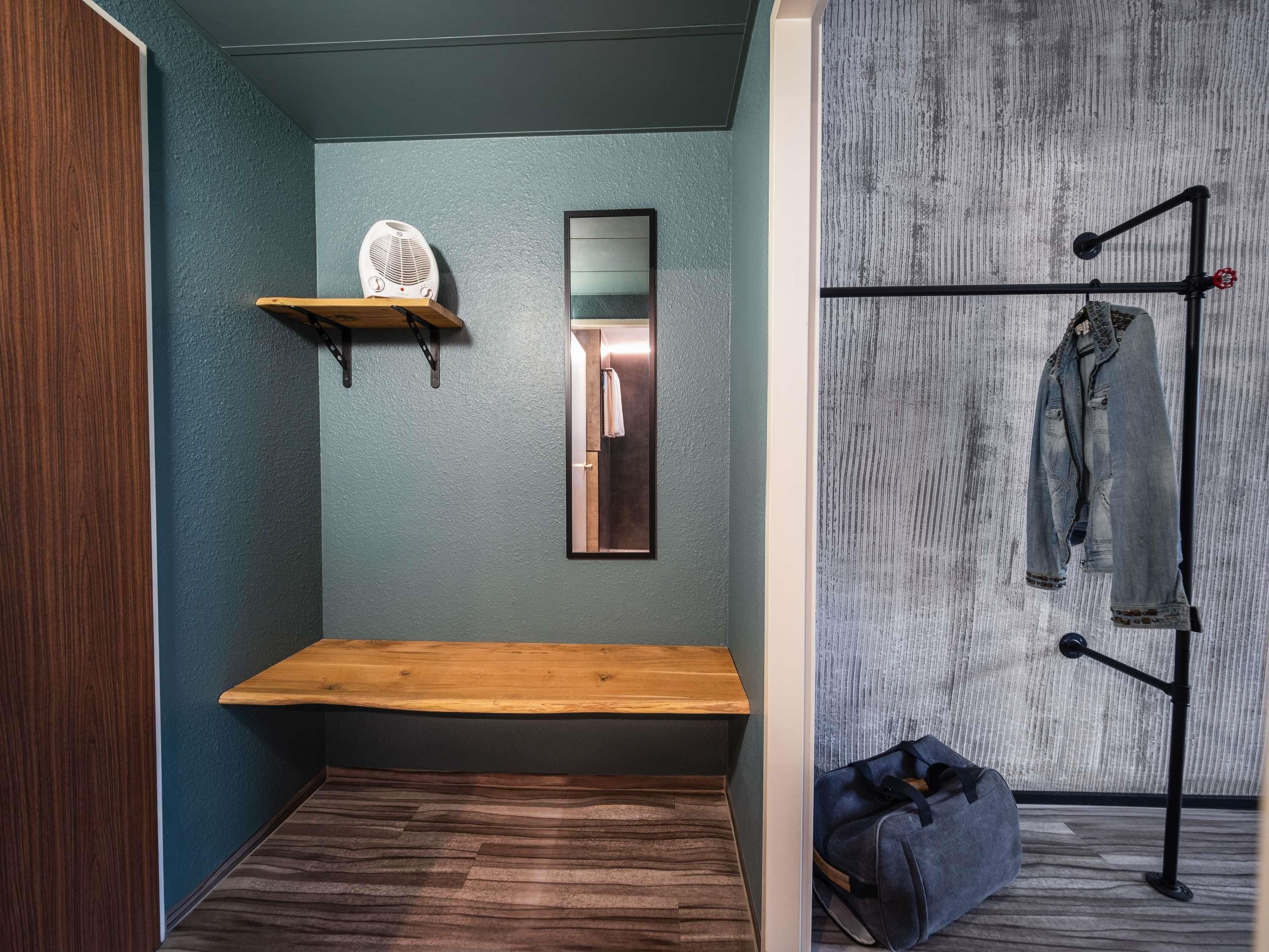 Una habitación pequeña con un banco de madera, un espejo, un perchero con una chaqueta y un bolso en el suelo.
