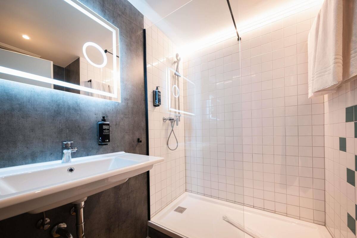 Uma casa de banho moderna com uma sanita branca, um espelho iluminado circularmente, um chuveiro com chuveiro de mão e um porta-toalhas.