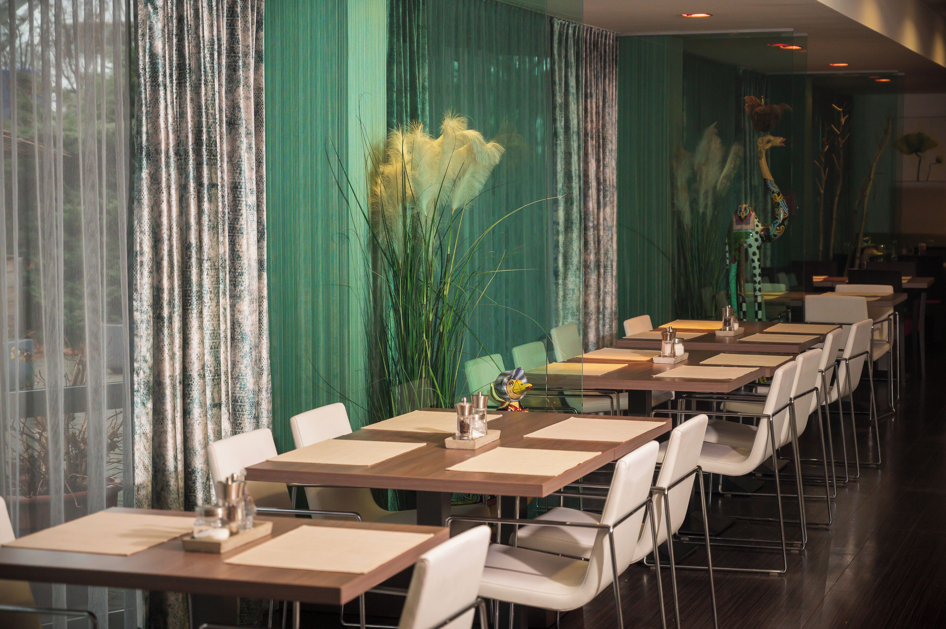 Ein Restaurant mit langen Tischen und weißen Stühlen, grünen Vorhängen und Zierpflanzen.
