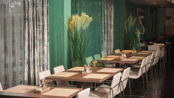 Ein Restaurant mit langen Tischen und weißen Stühlen, grünen Vorhängen und Zierpflanzen.