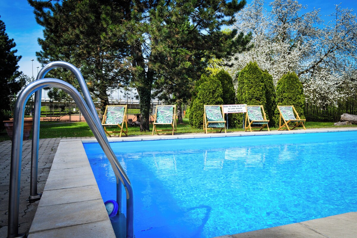 A swimming pool with a metal ladder, surrounded by trees and wooden chairs with signs.