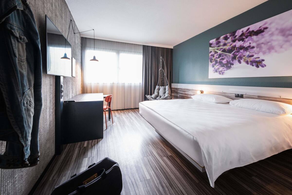 A modern hotel room with a large bed, a desk, a hanging chair, and a large window with curtains.