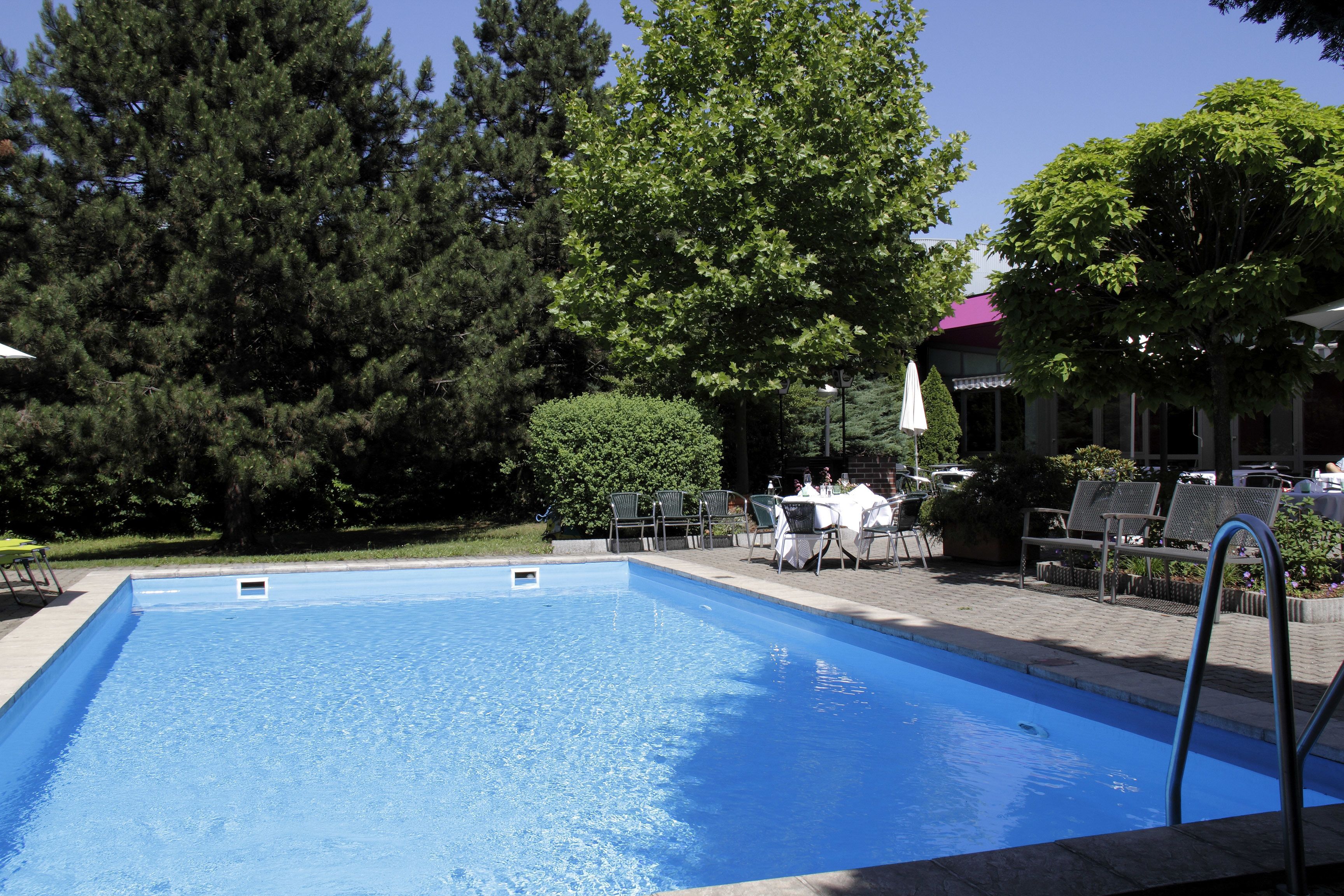 Una piscina rectangular con una escalera metálica en un lateral, rodeada por una zona pavimentada con mobiliario exterior y vegetación.