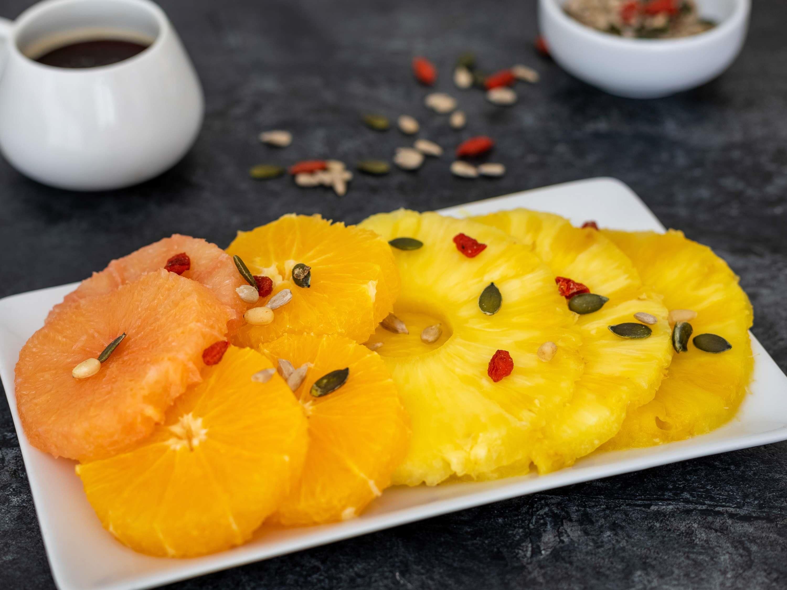 A plate of sliced oranges and pineapple with seeds and red berries, accompanied by small bowls of seeds and a cup of coffee.
