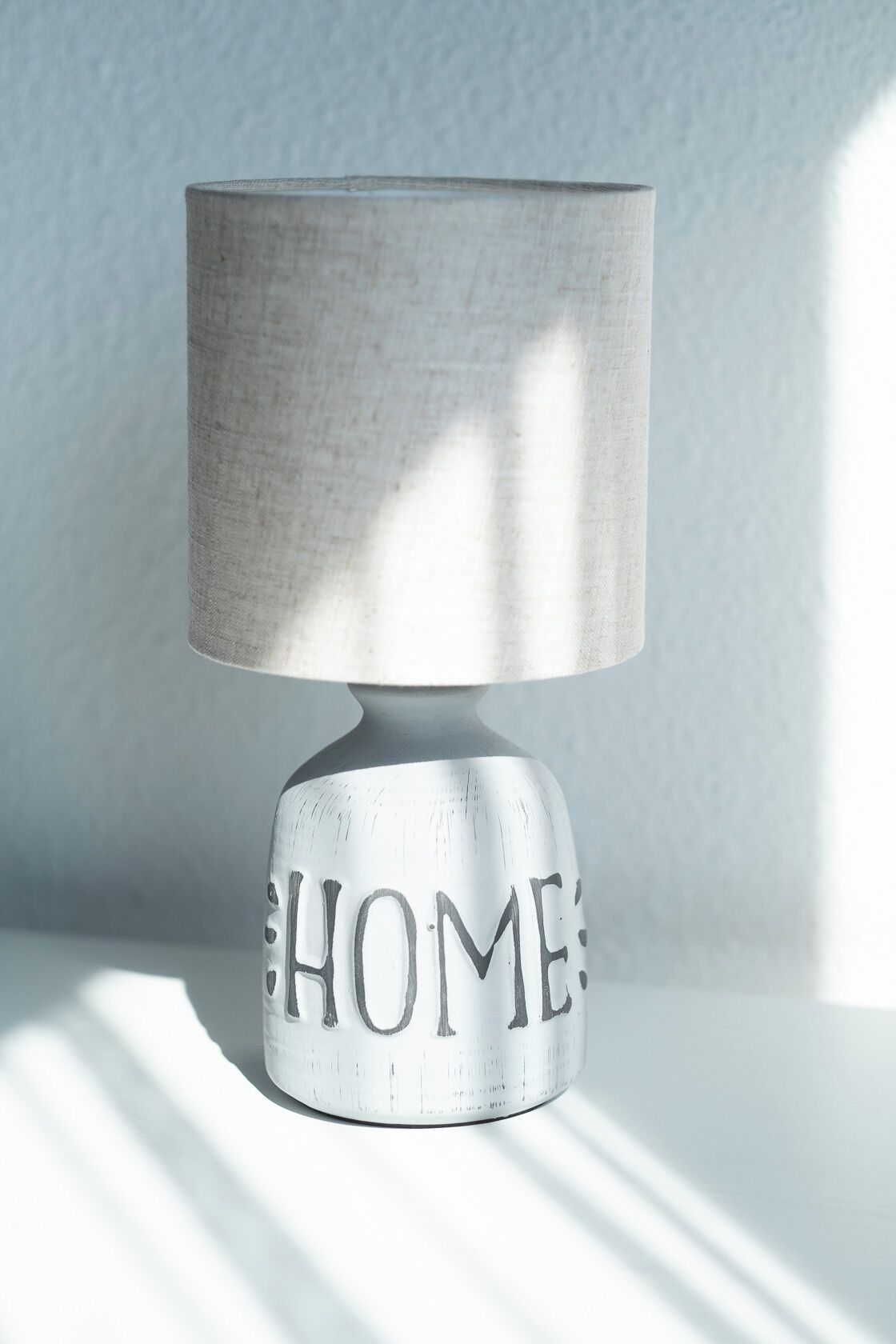 Una lámpara blanca con pantalla cilíndrica y una base con la palabra 'HOME' escrita.