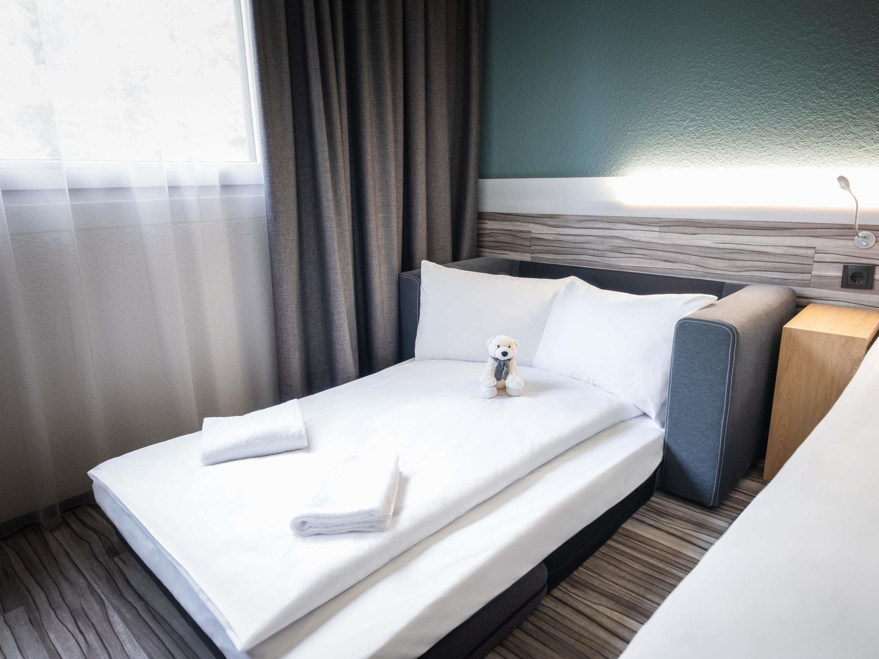A neatly made bed with white linens and two towels, a small teddy bear on the bed, a grey upholstered headboard, and a wooden nightstand with a lamp.