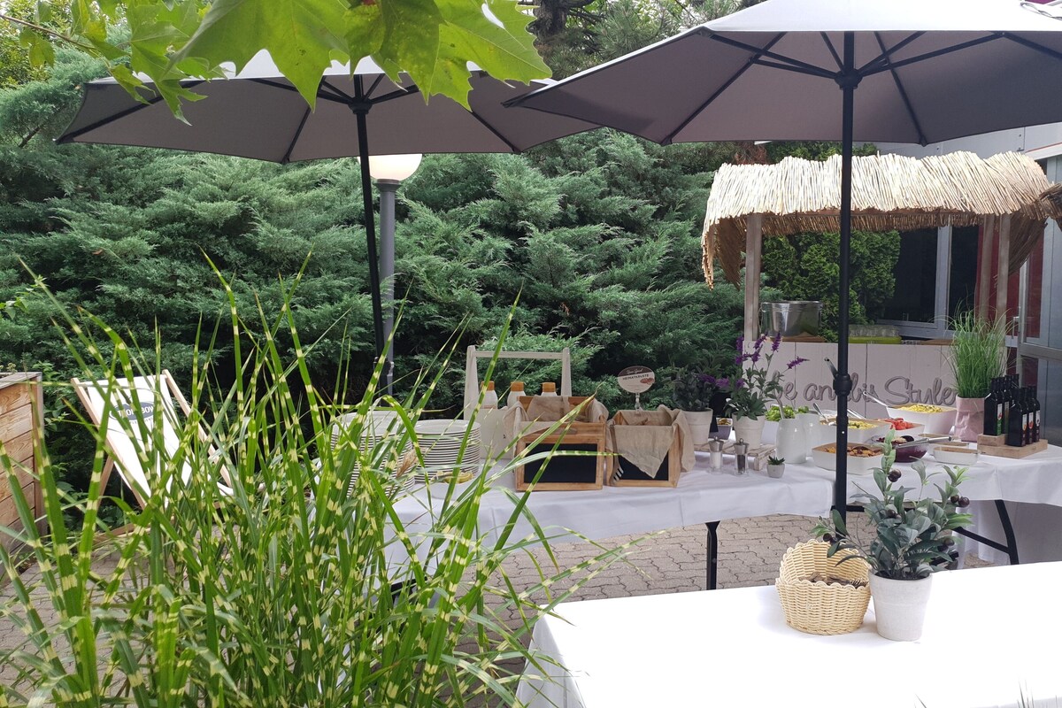 An outdoor dining area with tables covered by umbrellas, surrounded by greenery and potted plants.
