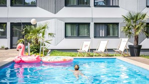 A swimming pool with a pink flamingo float and lounge chairs in front of a modern building with large windows. - ibis Styles Linz (Linz)