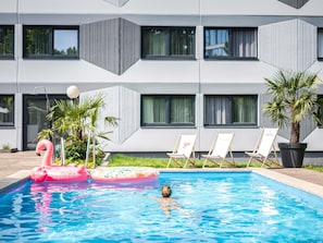 A swimming pool with a pink flamingo float and lounge chairs in front of a modern building with large windows. - ibis Styles Linz (Linz)