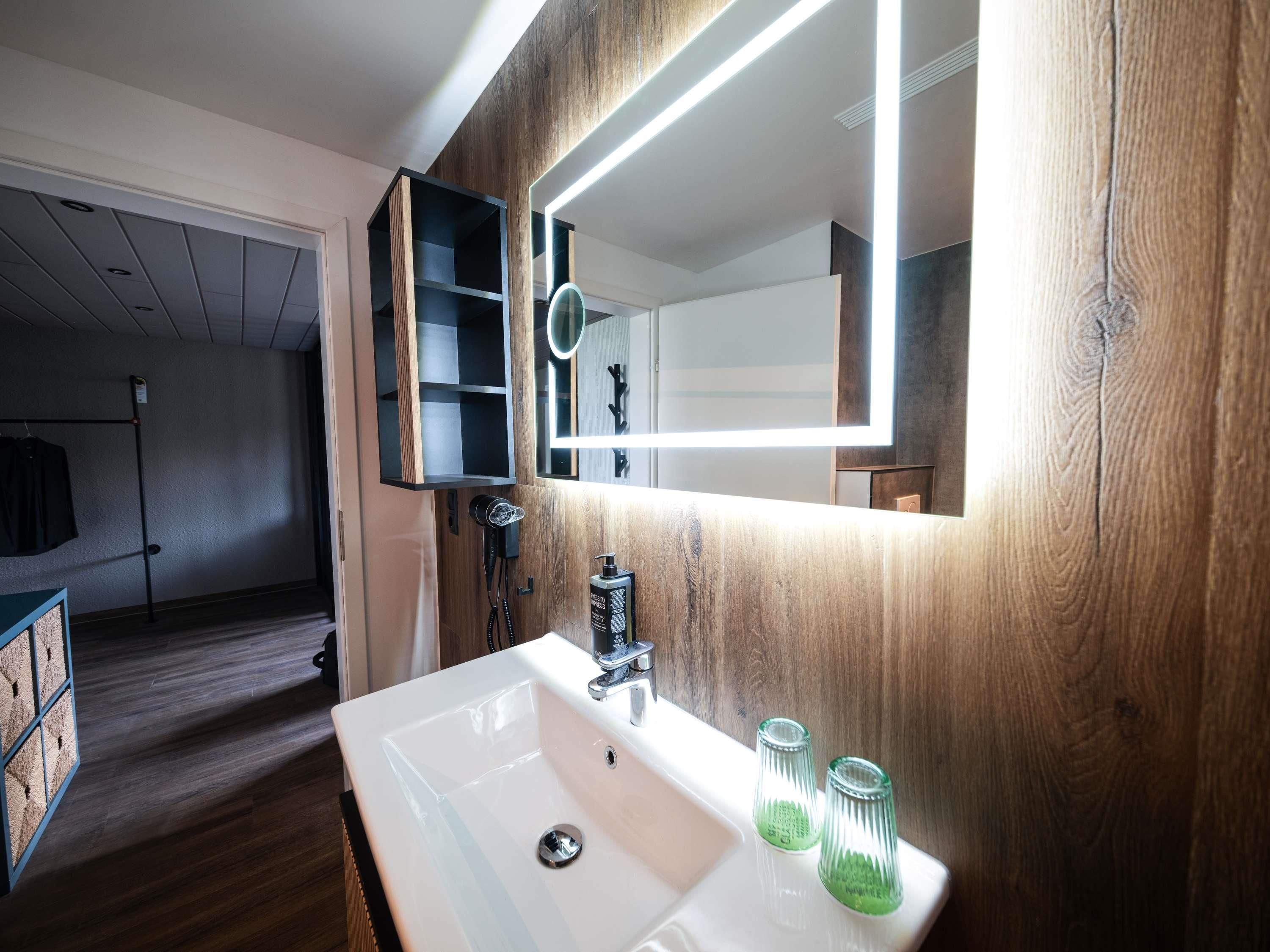 A modern bathroom with a white sink, wooden paneling, a mirror with integrated lighting, and a hairdryer.