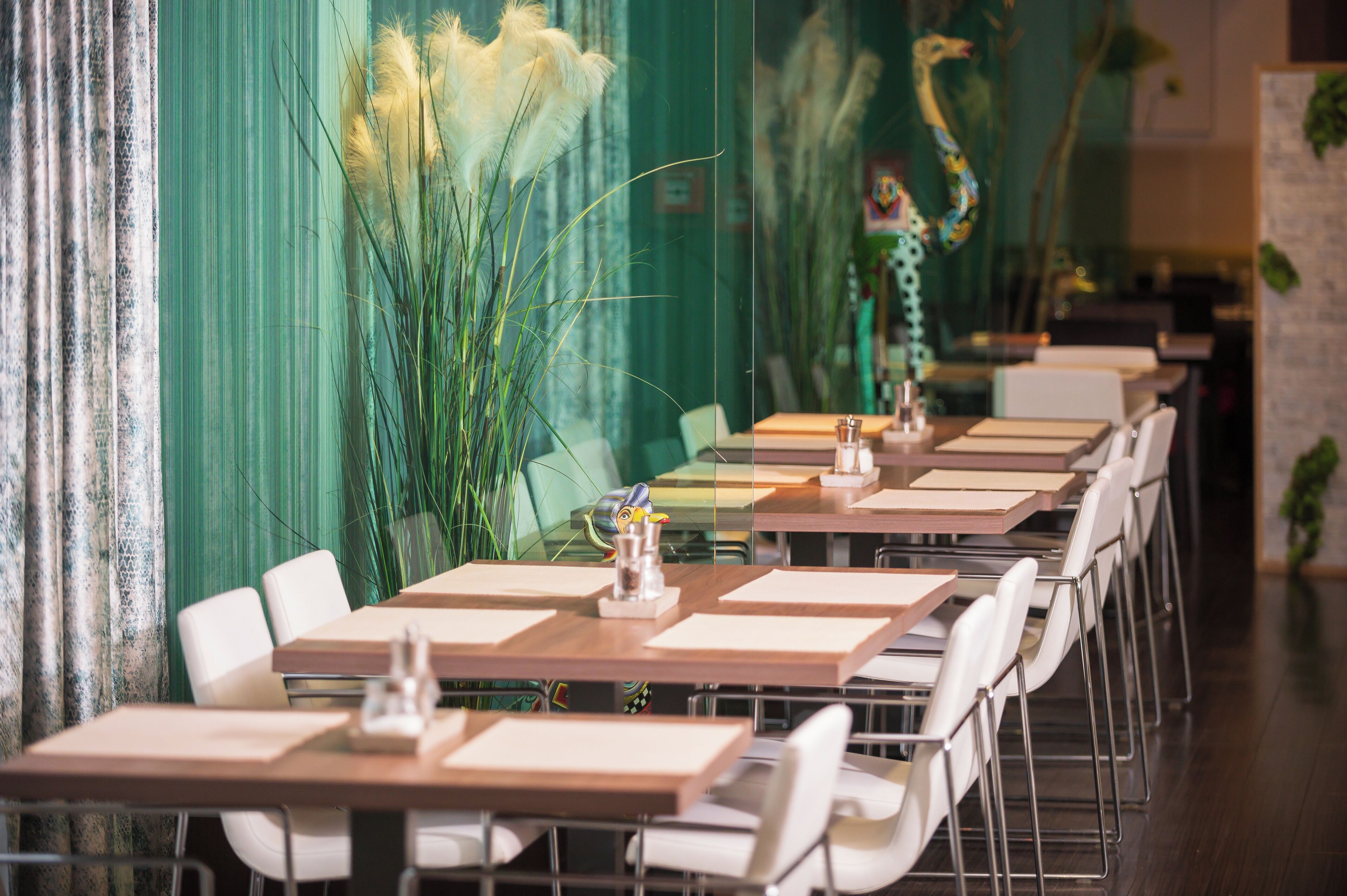 Un restaurante con mesas largas y sillas blancas, paredes verdes y plantas decorativas.
