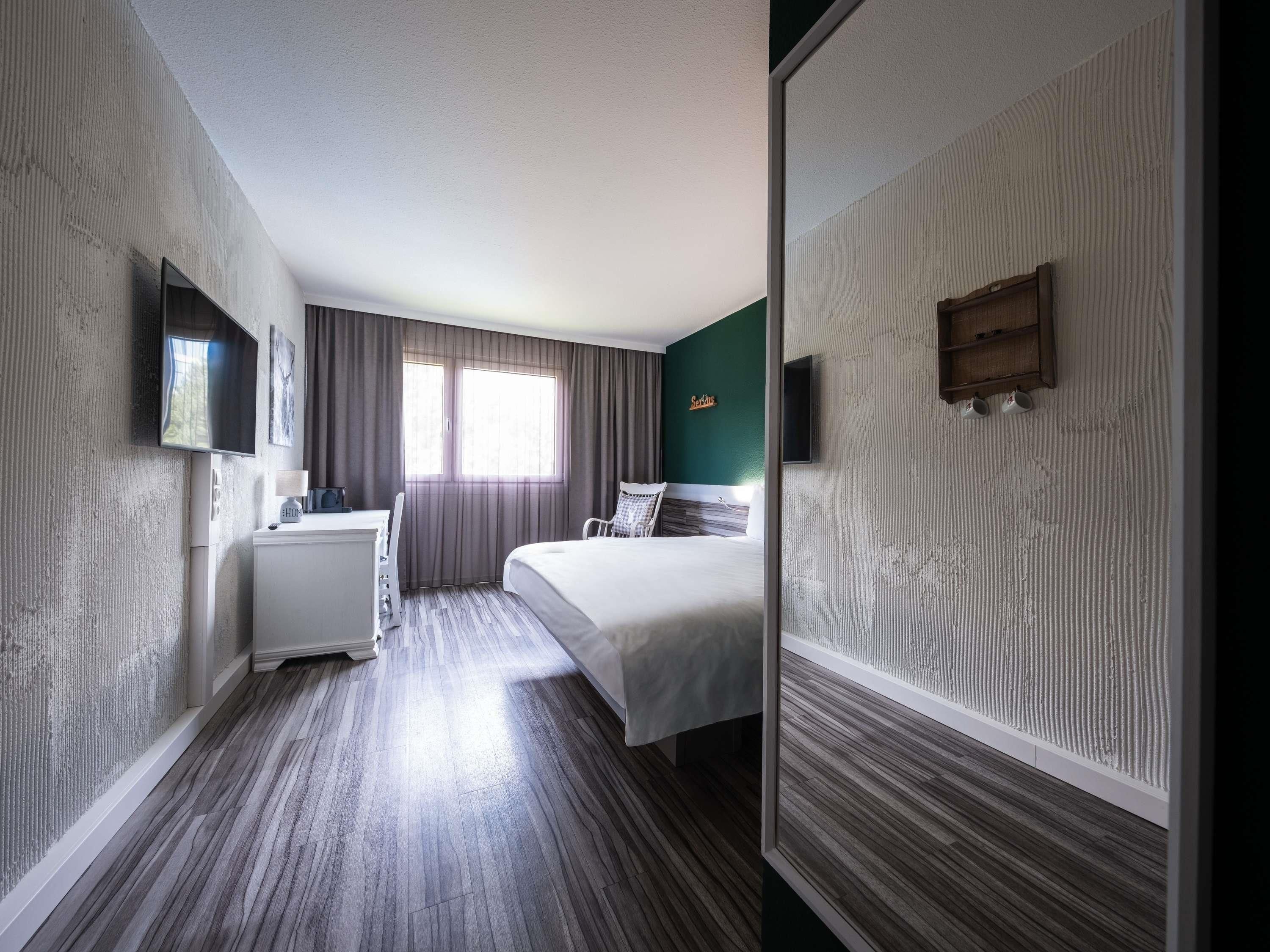 Une chambre d’hôtel avec un lit, un bureau, une chaise, une télévision et une fenêtre avec des rideaux.