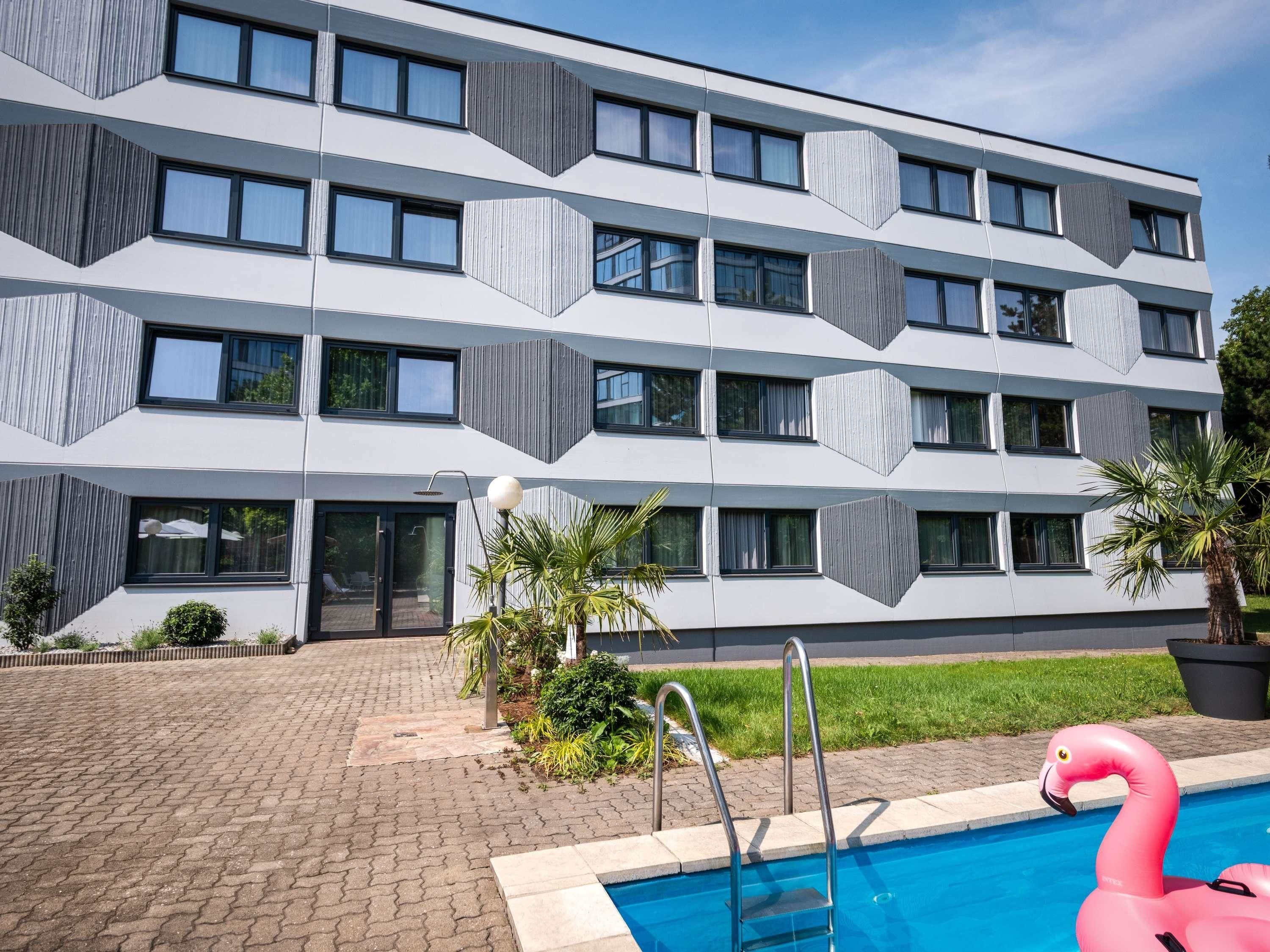 A modern hotel with a geometric facade, a swimming pool with a pink flamingo float, and a well-maintained garden area.