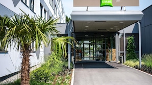 Una entrada de hotel moderna con fachada de vidrio, palmeras y un letrero que dice 'ibis Styles Linz Hotels'.