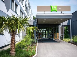 Una entrada de hotel moderna con fachada de vidrio, palmeras y un letrero que dice 'ibis Styles Linz Hotels'.