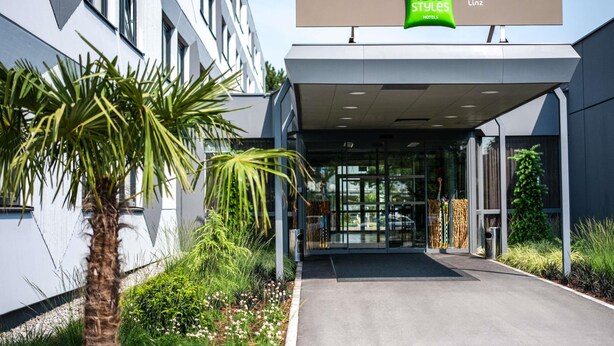 A modern hotel entrance with a glass facade, palm trees, and a sign for 'ibis Styles Linz Hotels.'
