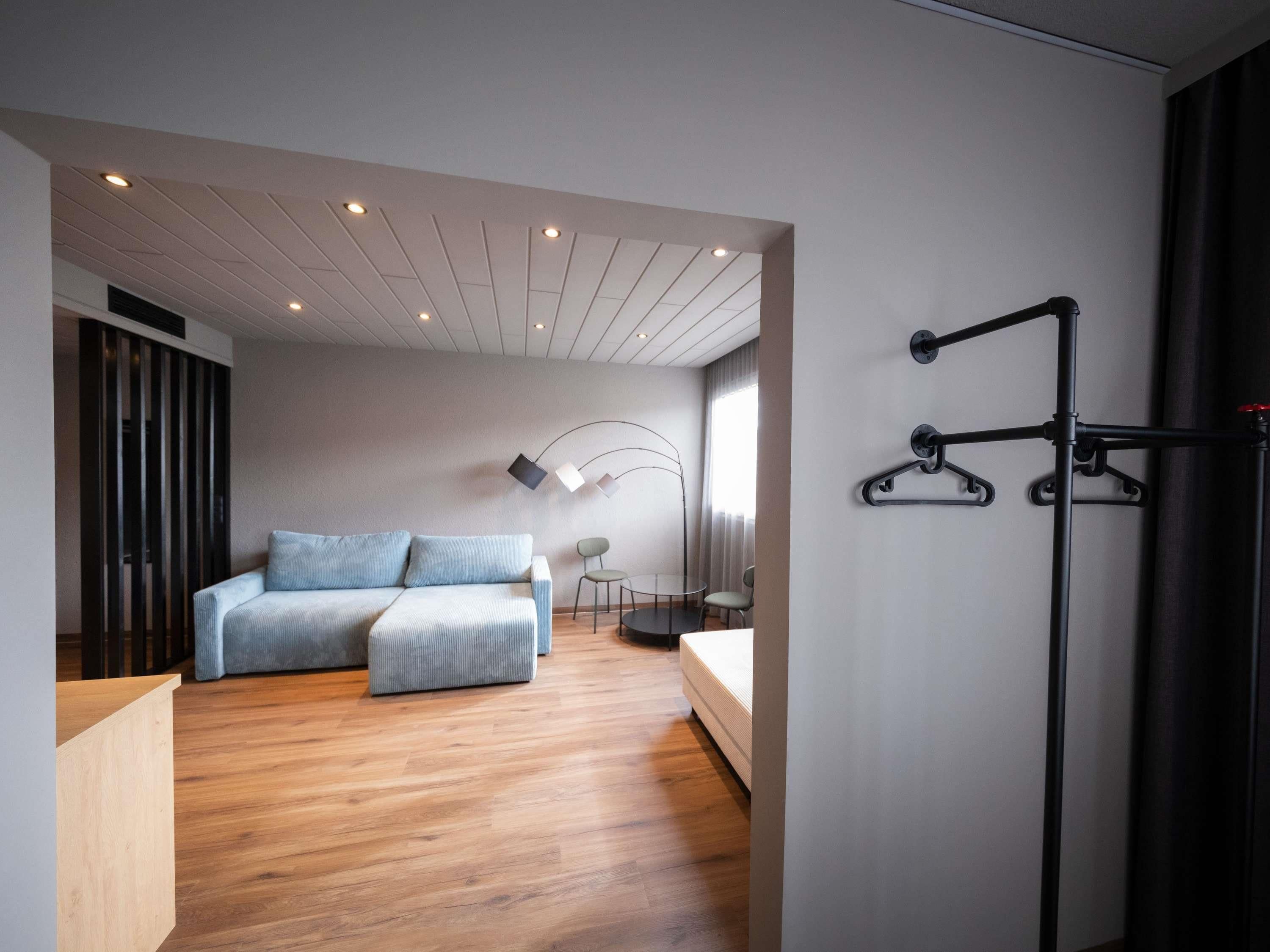 A modern hotel room with a wooden floor, a sofa, a bed, a desk, and a wall-mounted rack.