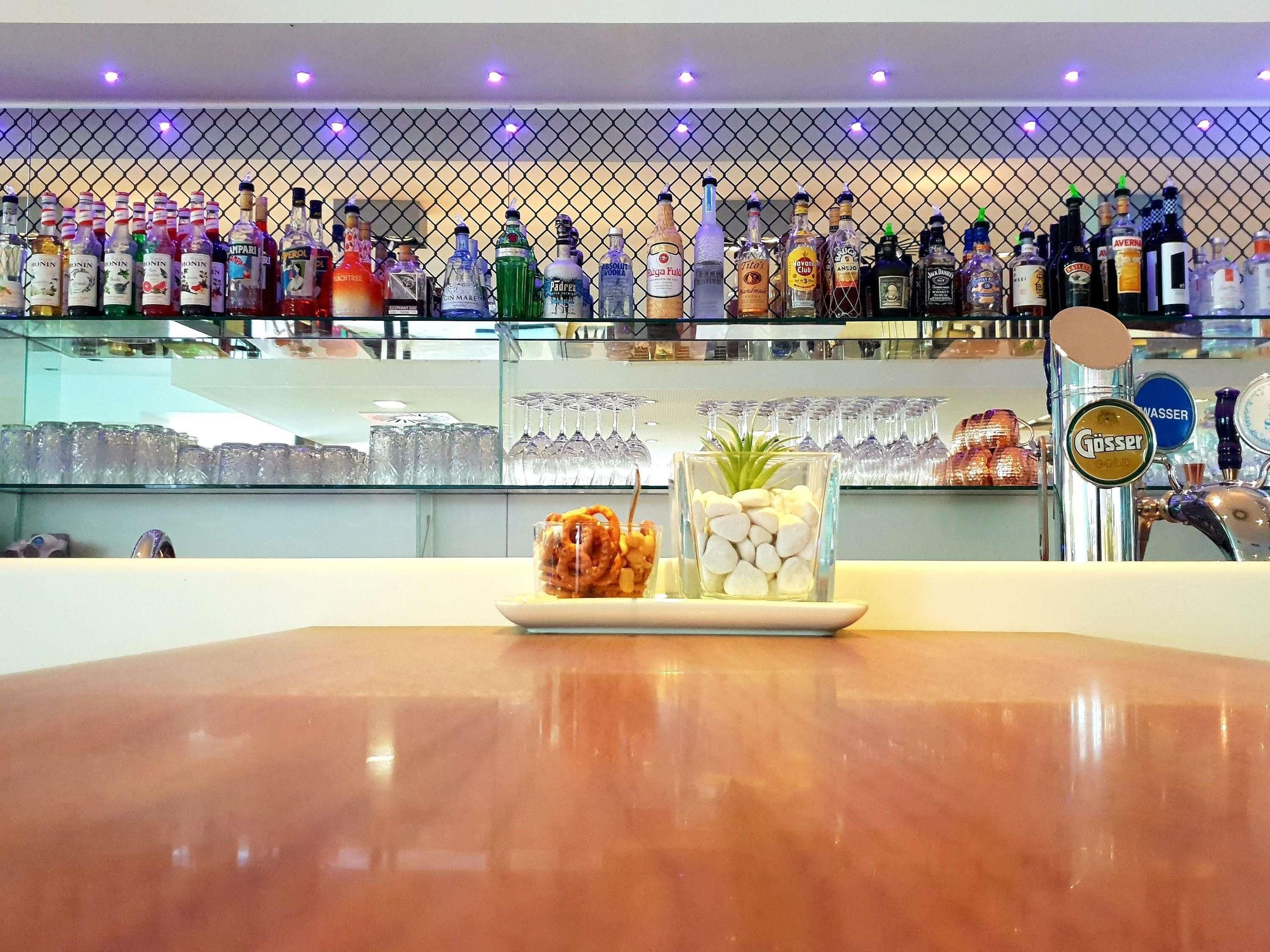 Zona de bar con una variedad de botellas de licor, una vitrina con vasos y una barra con un plato de comida y objetos decorativos.