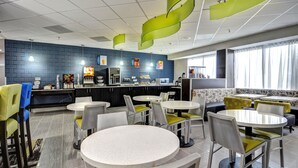 A modern dining area with round tables and chairs, a kitchen in the background, and a seating area with sofas and a coffee machine. - Best Western Plus Hanes Mall Hotel (Winston-Salem)