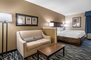 A hotel room with a sofa, a bed, a bedside table, a lamp, and framed artwork on the wall.