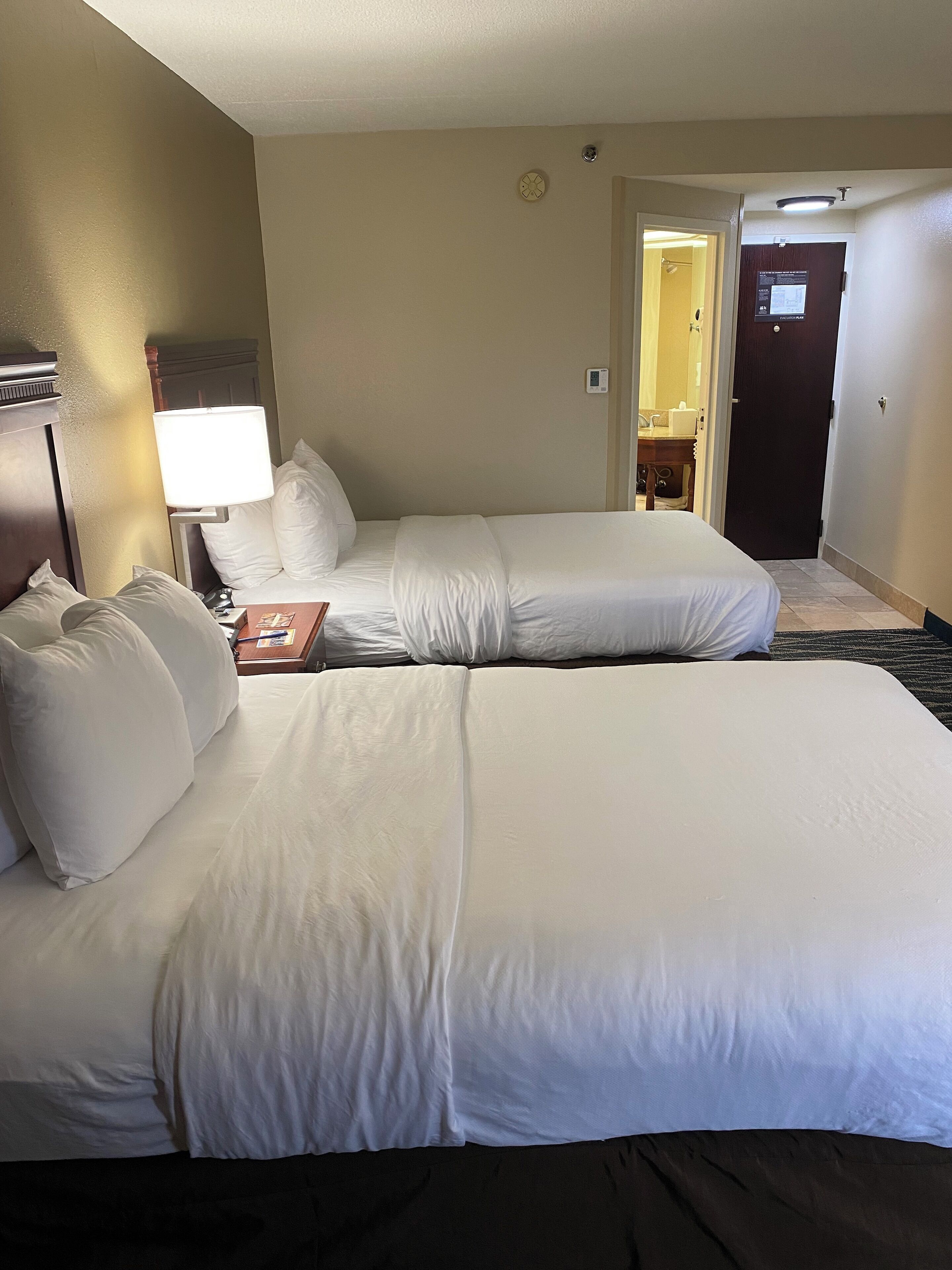 A hotel room with two beds, a nightstand, a lamp, and a door leading to a bathroom.